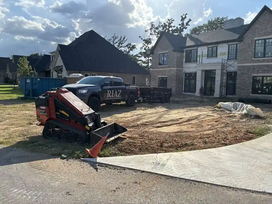 Orange stand-on skid steer grading a new-build lot beside a black pickup lettered RIAZ