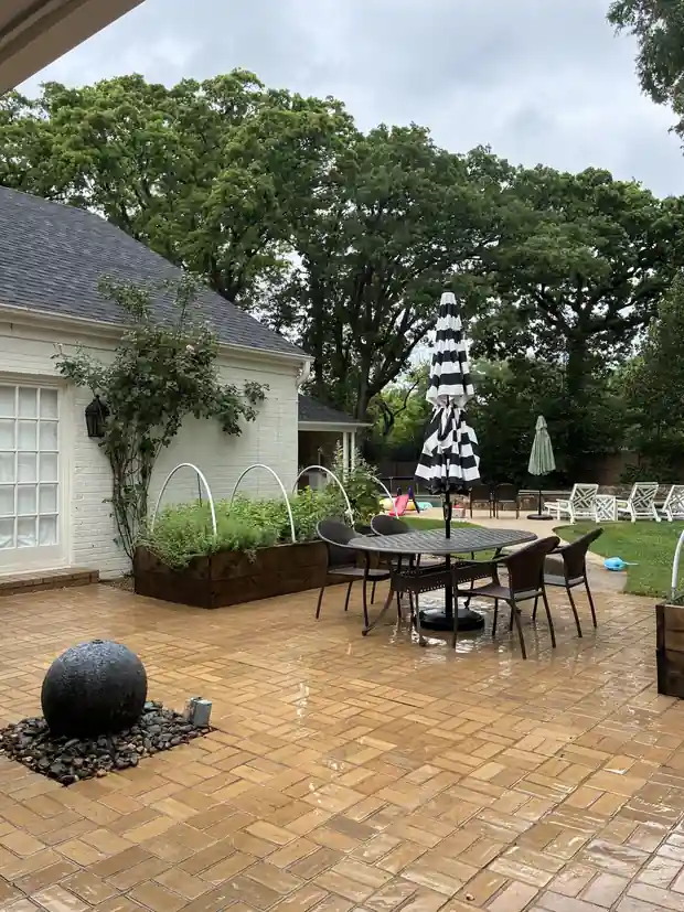 Wet basketball-weave paver patio with a black stone sphere fountain on river rock, cedar raised beds and an oak canopy behind a white brick house