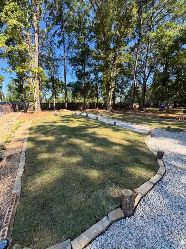 New sod strip edged by a natural stone border beside a gravel path, with pine woods behind