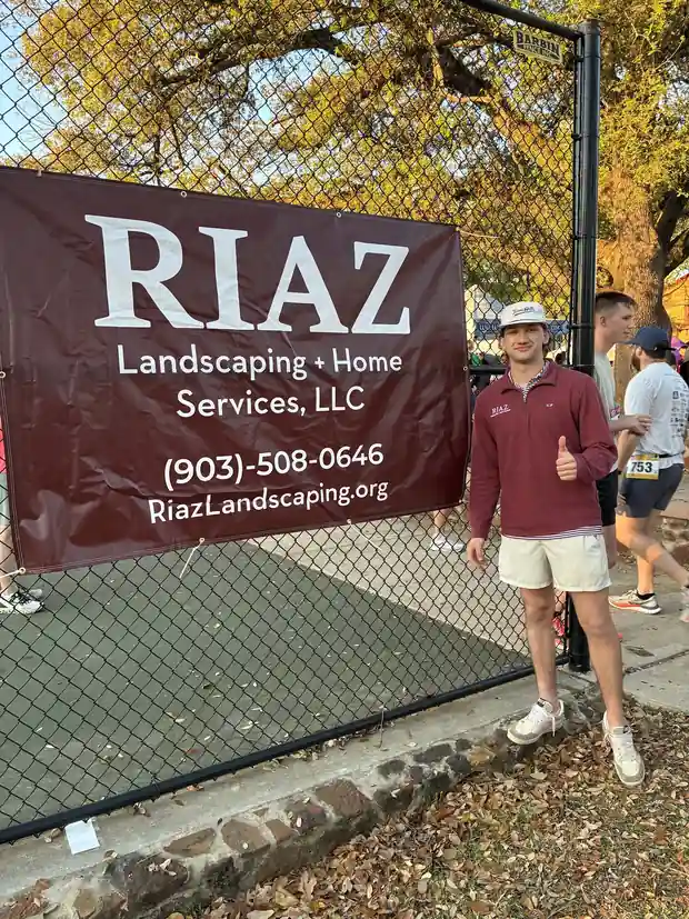 Carson Riaz giving a thumbs up beside the maroon RIAZ Landscaping + Home Services banner on a fence