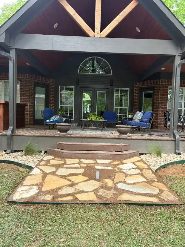 Tan flagstone pad at the steps of a timber-gable covered porch, framed by river rock beds and steel edging, a conduit stub in the stone