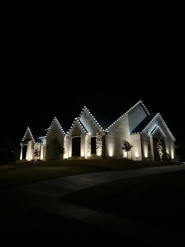 White roofline Christmas lights tracing every gable of a white brick home at night, with uplights on the walls