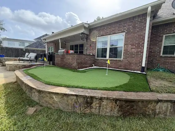 Artificial turf putting green with a flag on a raised stone-walled terrace behind a brick house