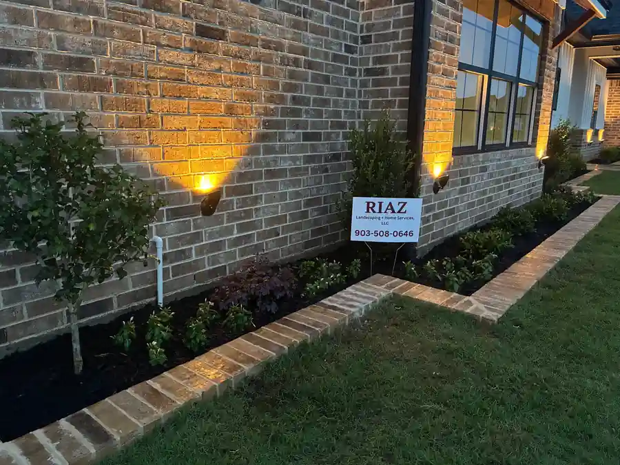 Warm uplights grazing a brick wall at dusk over a fresh mulch bed edged in brick pavers